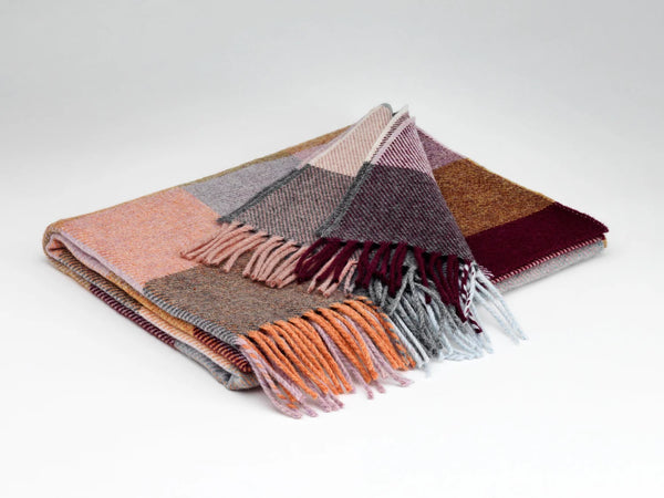 A folded plaid wool blanket with fringed edges, featuring a patchwork pattern of dusty pink, gray, brown, orange, and deep burgundy on a light gray background.