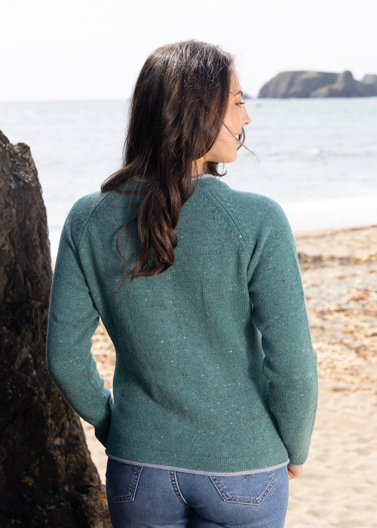 Woman wearing a green sweater standing on a beach looking out at the ocean.