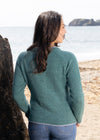 Woman wearing a green sweater standing on a beach looking out at the ocean.