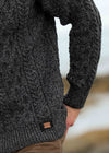 Close-up of a person wearing a dark charcoal gray cable-knit sweater, with the hand in a brown trouser pocket, against a blurred outdoor background. A small woven brand tag is visible near the hem, and the 'Aran Crafts' logo appears in the bottom right corner.