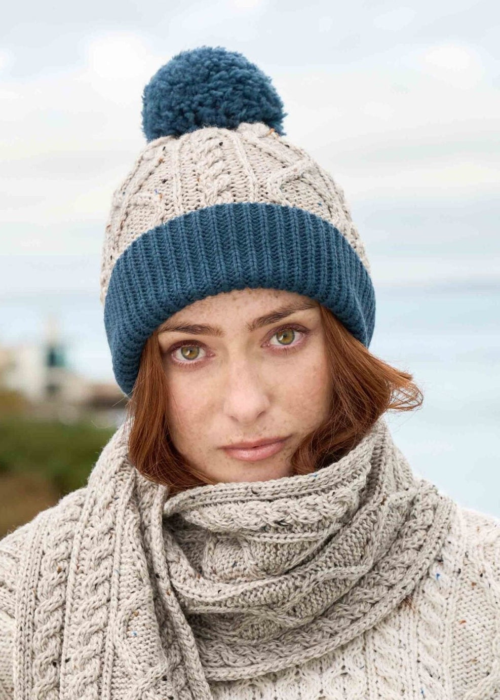 Woman wearing a cable knit sweater, scarf, and beanie with a bobble on a coastal background