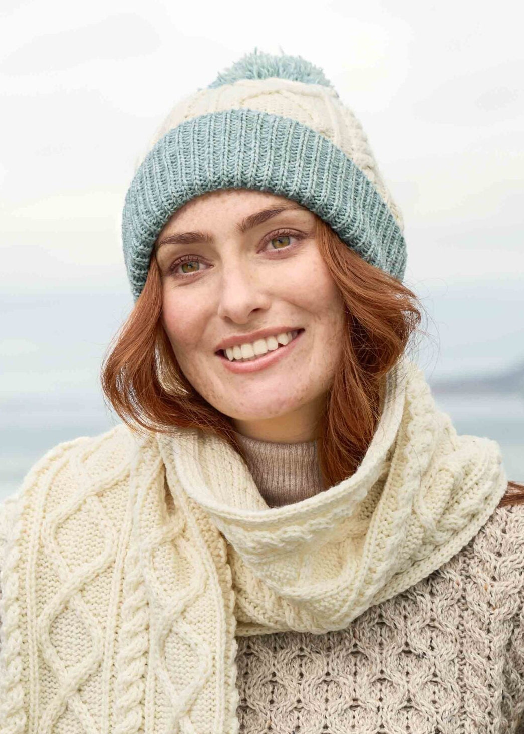 Woman wearing a cable knit sweater, scarf, and hat with a blurred natural background
