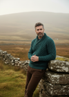 Man in a teal sweater standing by a stone wall with a misty landscape in the background
