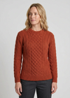 A woman wearing a rust-orange cable-knit crewneck sweater and dark gray trousers, standing against a light gray studio background.