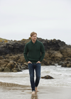 Man in a green sweater and jeans walking on a rocky beach.
