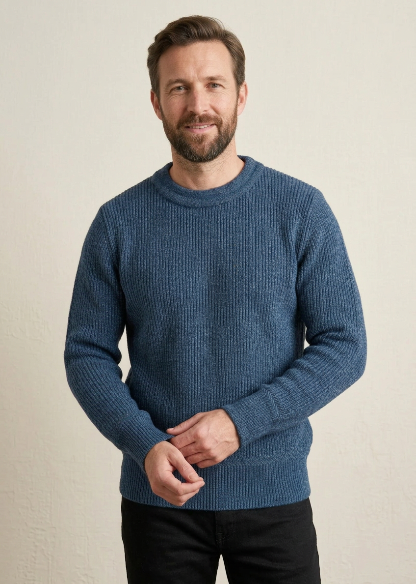 A man wearing a textured blue crewneck sweater, standing against a plain light beige wall.