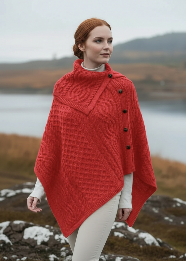 Woman wearing a red aran knitted poncho standing in a scenic outdoor setting with water and mountains.