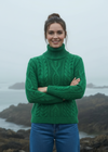 Woman wearing a green cable knit sweater standing by a body of water with mountains in the background.