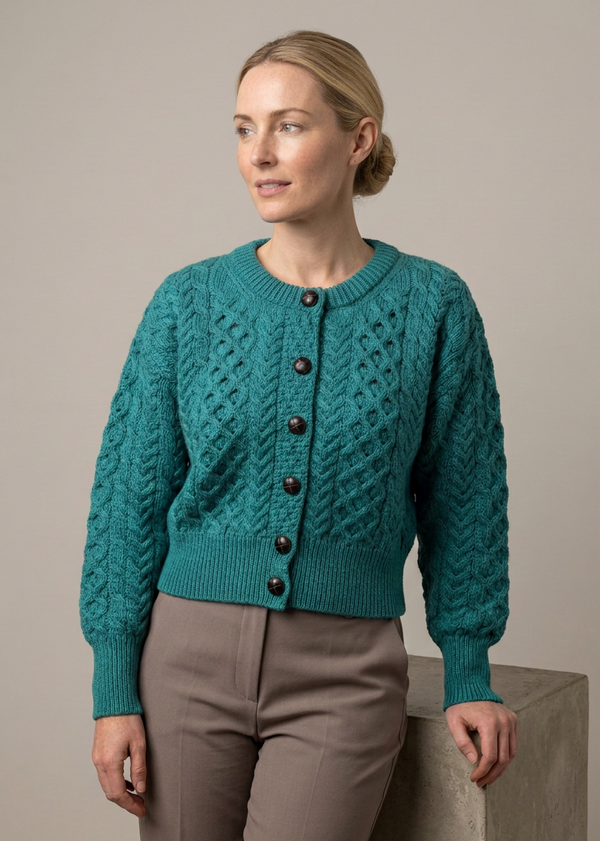 A woman wearing a teal cable-knit cropped cardigan sweater with dark buttons, paired with taupe trousers, standing next to a concrete block against a neutral beige background.