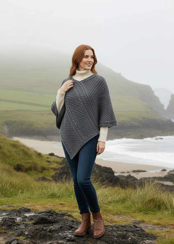 Woman wearing a gray knitted poncho standing in a scenic coastal area with misty mountains.