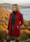 Woman wearing a red knitted coat standing in a scenic autumn landscape with trees and water.