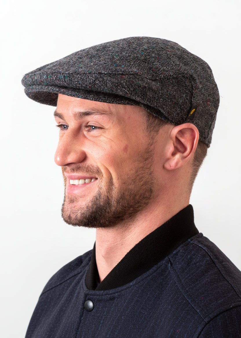 A man in profile wearing a dark gray tweed flat cap against a plain white background. HATMAN