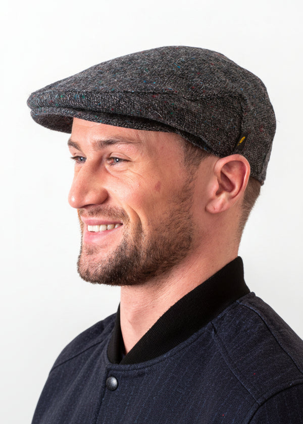 A man in profile wearing a dark gray tweed flat cap against a plain white background. HATMAN